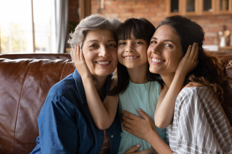 Head shot portrait happy three generations of women hugging, touching cheeks, smiling little girl sitting on couch between young mother and mature grandmother, posing for family photo at home